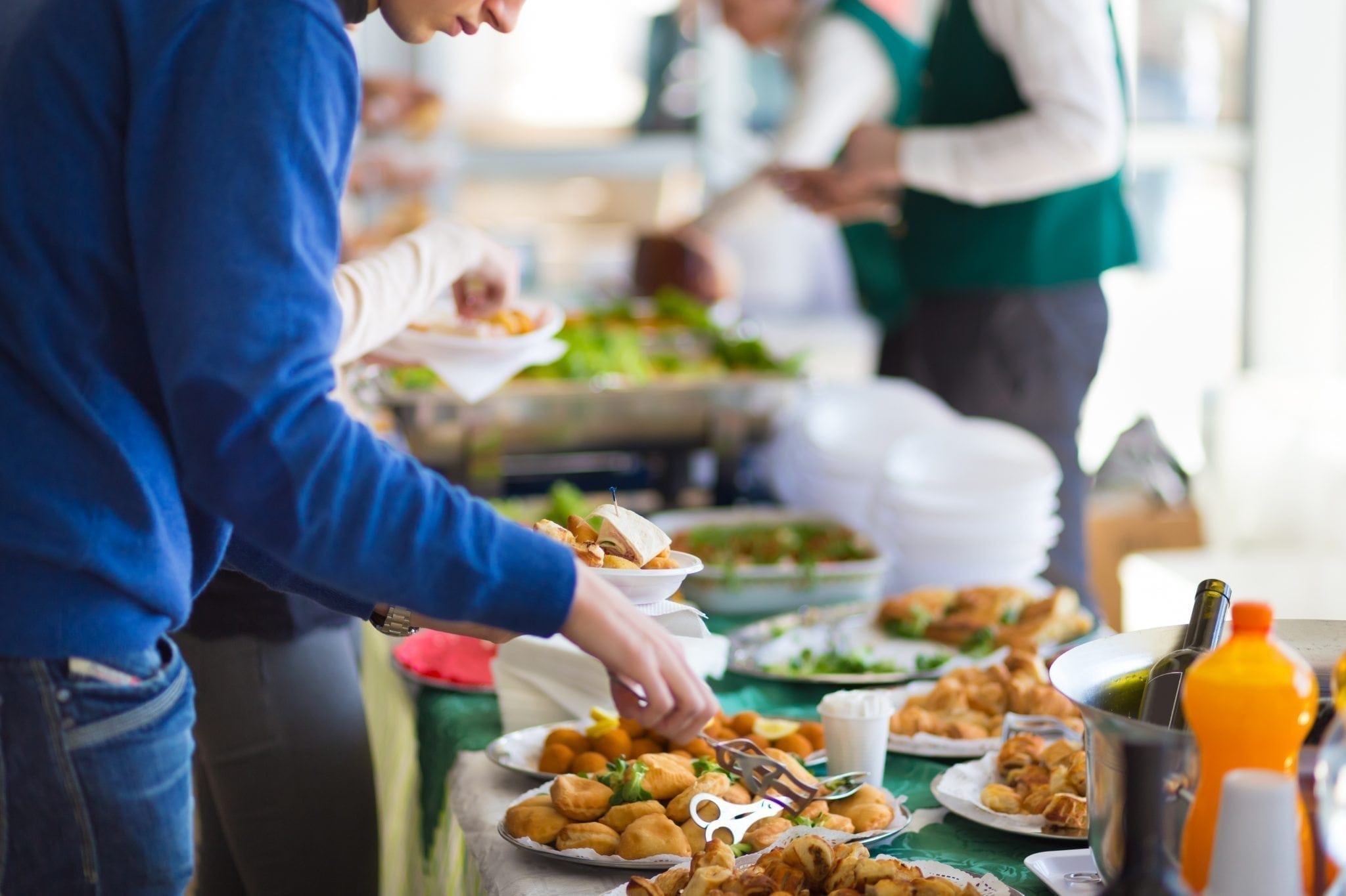 People are gathered around a buffet table selecting various foods. Plates of appetizers, main dishes, and bowls of salads are visible, along with disposable plates stacked at one end. Some individuals are serving themselves while others wait their turn, mindful of useful buffet tips for an efficient experience. MyFitnessPal Blog