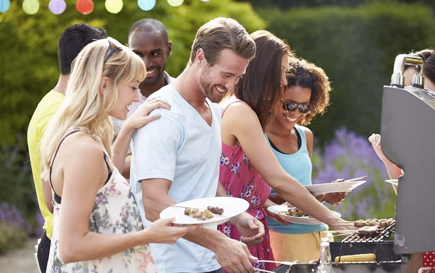A group of people gathers around a BBQ grill outdoors. They are smiling and holding plates, preparing and serving food, sharing joy in communal meals. Colorful lights are strung above, and there is greenery in the background. The scene suggests a casual, social gathering where emotional eating creates bonds. MyFitnessPal Blog