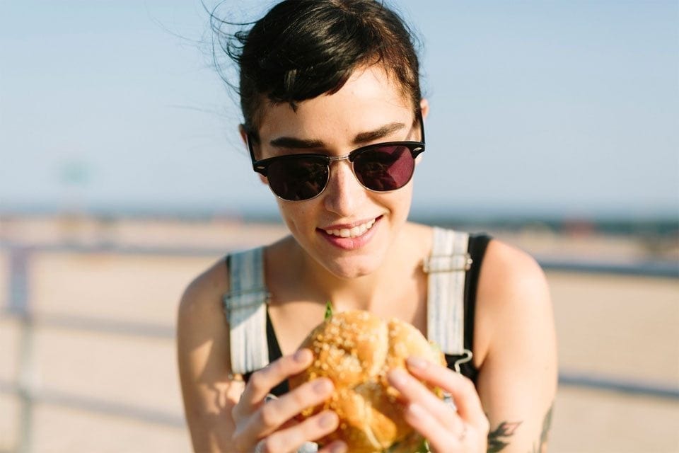 A person wearing sunglasses and a sleeveless top is holding a sandwich with both hands. The background is a sunny, blurred outdoor setting, possibly a beach. Smiling down at the sandwich, they appear to take control of their food choices, savoring each bite with satisfaction and joy. MyFitnessPal Blog