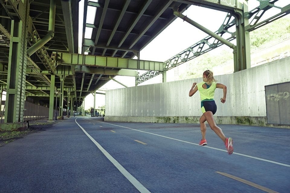 A person wearing a neon green top and black shorts is doing cardio by running on a paved road under a metal bridge. The scene appears to be in an urban-industrial area, with the bridge structure and concrete walls creating a geometric backdrop. The runner is facing away from the camera. MyFitnessPal Blog
