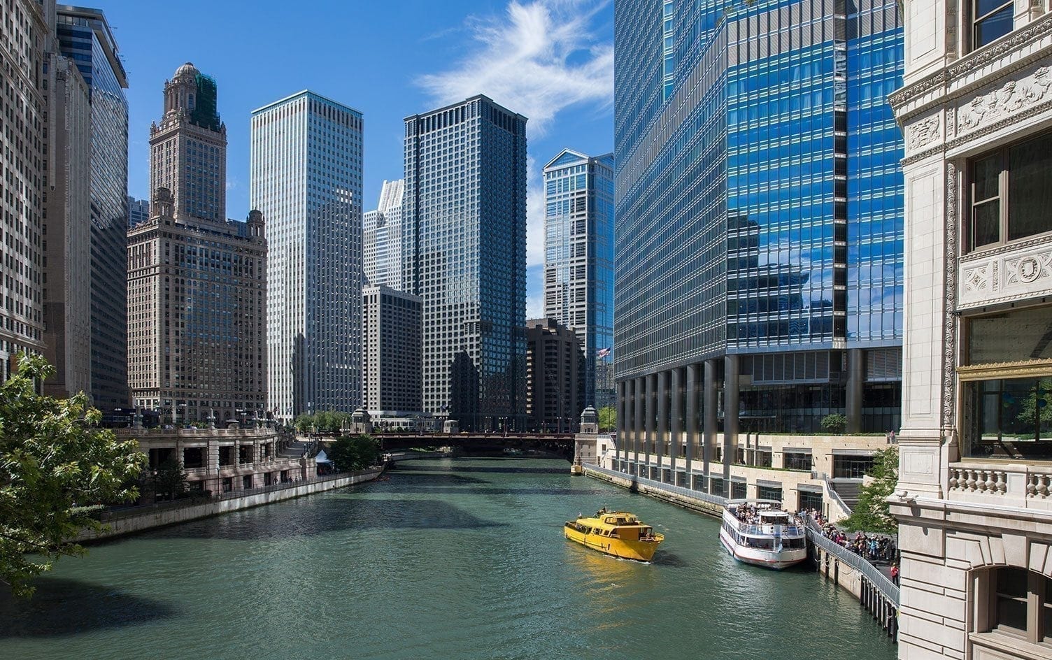 A view of a city river in Chicago surrounded by tall buildings on a sunny day. A yellow water taxi and a white boat are on the river, with people walking along the riverbank. The water appears green, and the sky is blue with scattered clouds. MyFitnessPal Blog