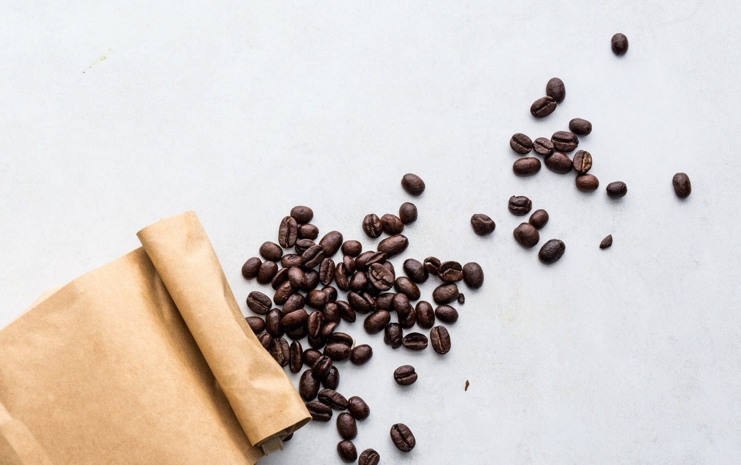 A brown paper bag tipped over on a white surface with coffee beans spilling out, fueling trends in minimalist kitchen aesthetics. The coffee beans are scattered in various positions on the surface around the bag. MyFitnessPal Blog