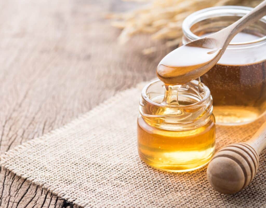 A small glass jar filled with honey sits on a piece of burlap cloth on a wooden surface. A wooden spoon drizzles honey into the jar, showcasing a dietitian’s easy pre-workout hack. Another larger jar of honey is visible in the background along with a honey dipper. What a Dietitian Says About A Viral Pre-workout Hack MyFitnessPal Blog